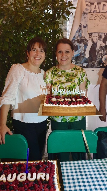 Due donne sorridenti reggono insieme una torta decorata con frutti di bosco e scritta di auguri
