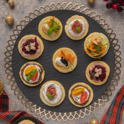 Blinis assortiti con salmone, gamberi, prosciutto crudo, robiola e pomodorini disposti su vassoio natalizio