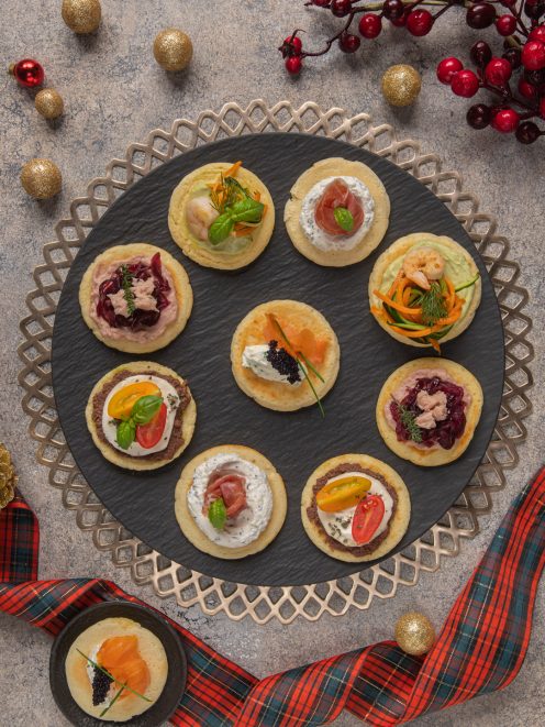 Blinis assortiti con salmone, gamberi, prosciutto crudo, robiola e pomodorini disposti su vassoio natalizio