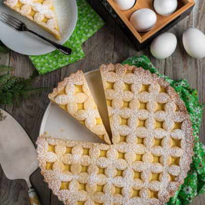Crostata con crema pasticcera decorata con reticolo di pasta frolla e zucchero a velo, una fetta servita a parte