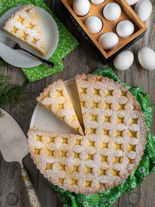 Crostata con crema pasticcera decorata con reticolo di pasta frolla e zucchero a velo, una fetta servita a parte