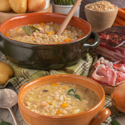 Gerstsuppe servita in due ciotole di terracotta con orzo, verdure e speck a vista