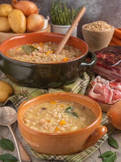 Gerstsuppe servita in due ciotole di terracotta con orzo, verdure e speck a vista
