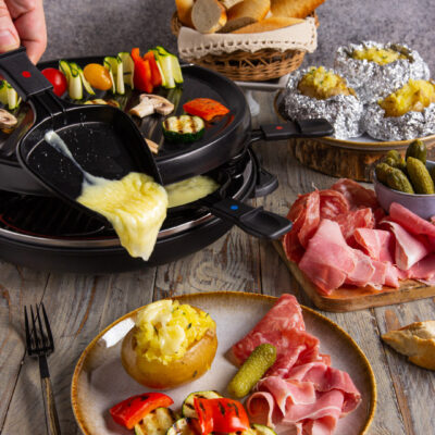 Raclette con formaggio filante versato su patata, verdure grigliate e affettati