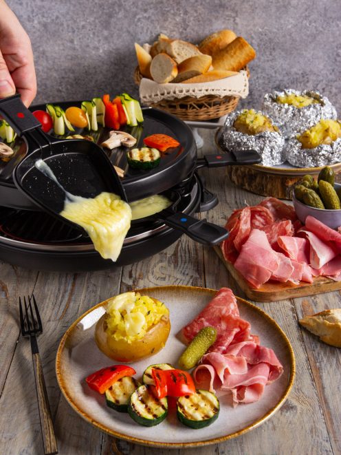 Raclette con formaggio filante versato su patata, verdure grigliate e affettati
