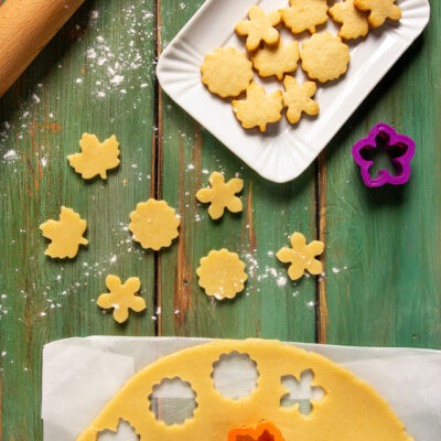 Pasta frolla senza uova stesa con biscotti ritagliati a forma di fiore e foglia, crudi e cotti