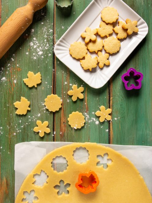 Pasta frolla senza uova stesa con biscotti ritagliati a forma di fiore e foglia, crudi e cotti