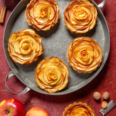 Crostatine di mele con fette disposte a rosa su vassoio in metallo, mele fresche e cannella accanto