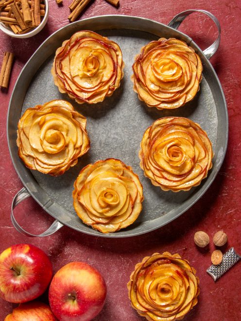 Crostatine di mele con fette disposte a rosa su vassoio in metallo, mele fresche e cannella accanto