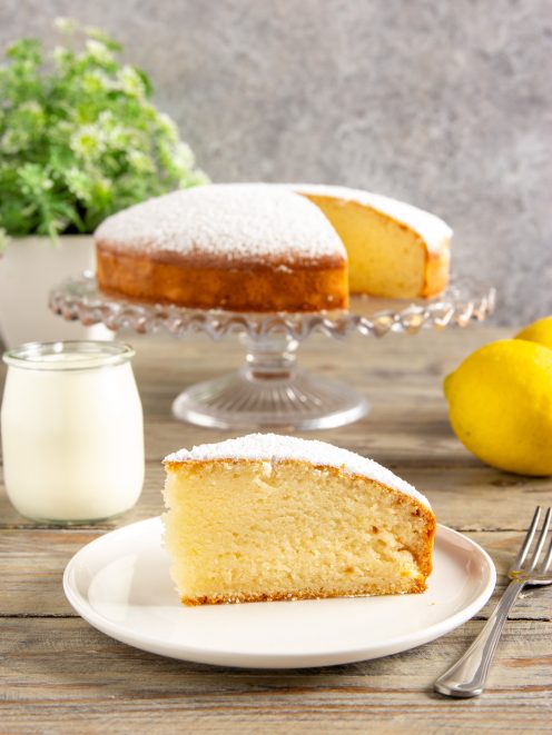 Torta allo yogurt senza uova a fette, soffice e spolverata di zucchero a velo, con limoni freschi accanto
