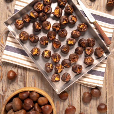 Castagne al forno con incisione a croce su teglia, affiancate da castagne crude