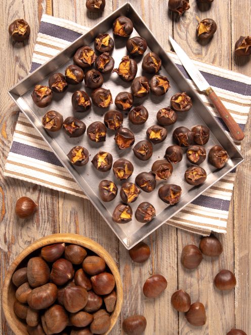Castagne al forno con incisione a croce su teglia, affiancate da castagne crude