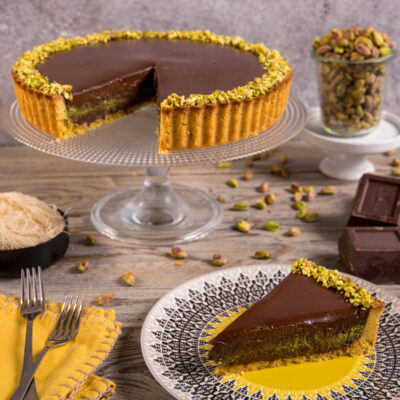 Crostata Dubai al cioccolato e pistacchio con una fetta servita, bordo decorato con granella di pistacchi