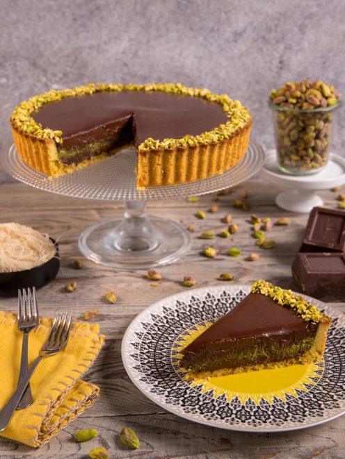 Crostata Dubai al cioccolato e pistacchio con una fetta servita, bordo decorato con granella di pistacchi