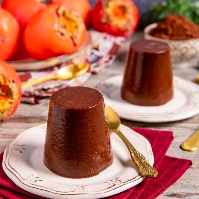 Budino di cachi e cacao sformato, con superficie lucida e compatta, servito con cachi maturi sullo sfondo