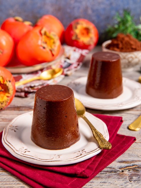 Budino di cachi e cacao sformato, con superficie lucida e compatta, servito con cachi maturi sullo sfondo