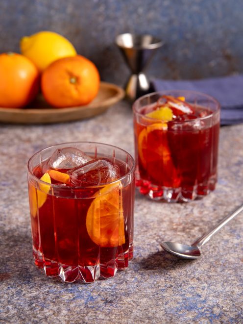 Americano cocktail servito in due bicchieri bassi con fetta di arancia, scorza di limone e cubetti di ghiaccio