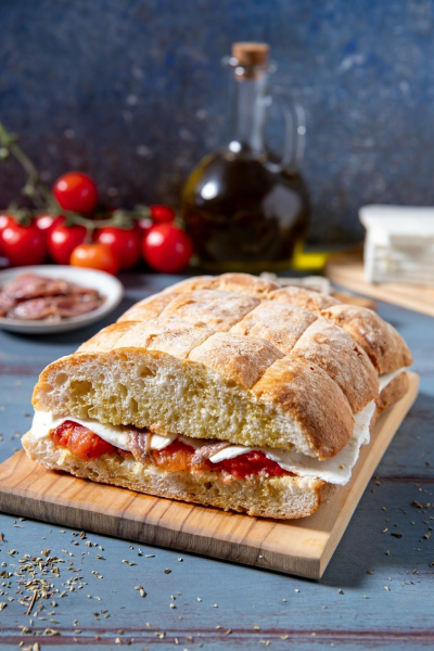 Pane cunzato farcito con pomodori, mozzarella e acciughe, servito su tagliere di legno