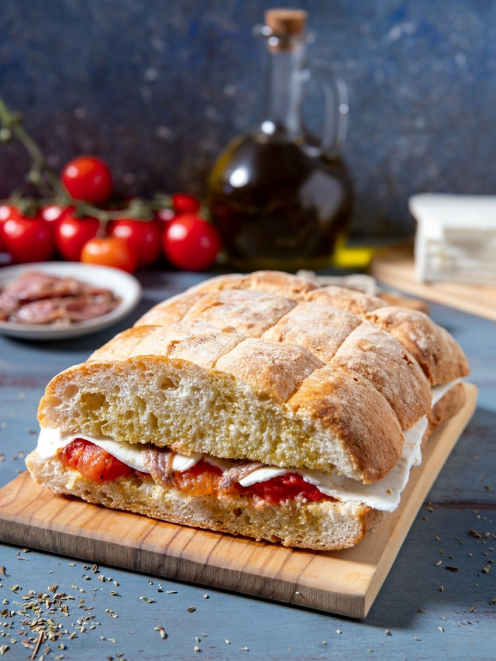 Pane cunzato farcito con pomodori, mozzarella e acciughe, servito su tagliere di legno