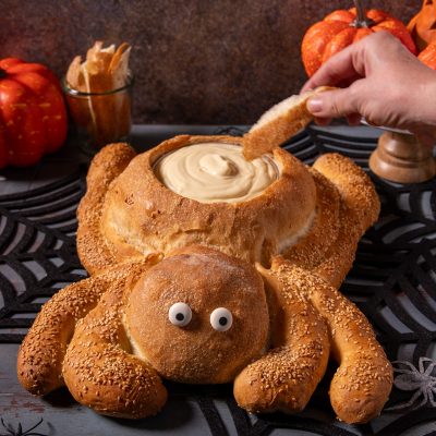 Pane ragno con zampe ricoperte di semi di sesamo, occhi decorativi e cavità ripiena di crema, mentre una mano intinge un pezzo di pane