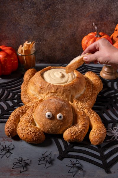 Pane ragno con zampe ricoperte di semi di sesamo, occhi decorativi e cavità ripiena di crema, mentre una mano intinge un pezzo di pane