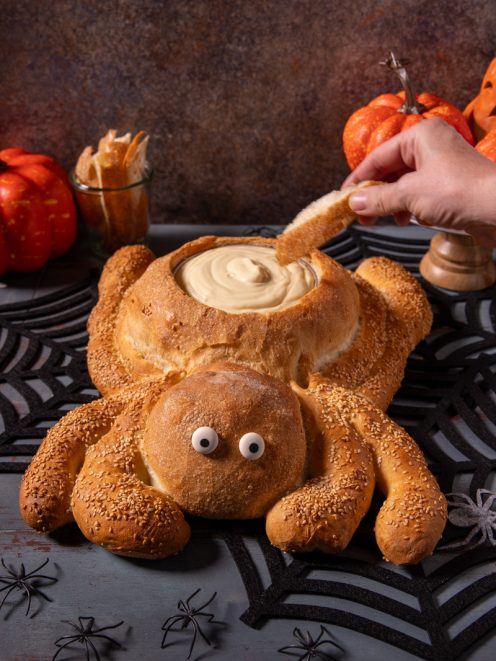 Pane ragno con zampe ricoperte di semi di sesamo, occhi decorativi e cavità ripiena di crema, mentre una mano intinge un pezzo di pane
