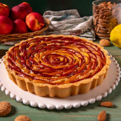 Crostata di pesche e amaretti con fettine di pesca disposte a spirale su base di frolla e crema, servita intera