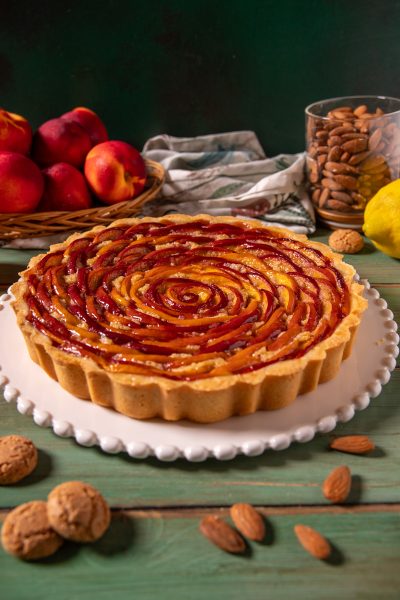 Crostata di pesche e amaretti con fettine di pesca disposte a spirale su base di frolla e crema, servita intera