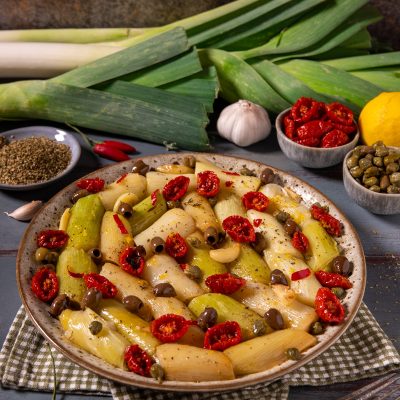 Porri alla mediterranea a pezzi con pomodori secchi, olive, capperi e spicchi d'aglio, serviti in un piatto da portata