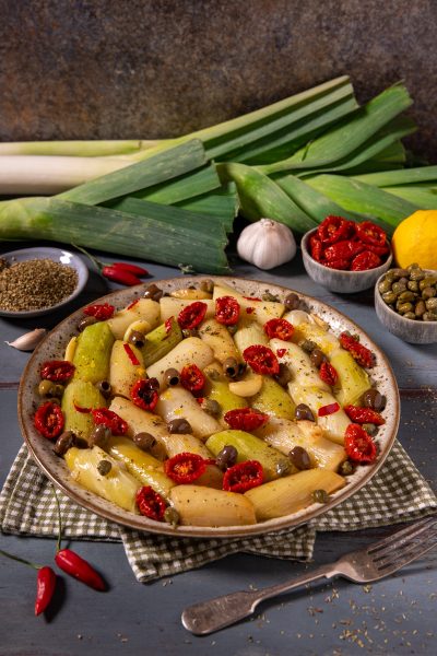 Porri alla mediterranea a pezzi con pomodori secchi, olive, capperi e spicchi d'aglio, serviti in un piatto da portata