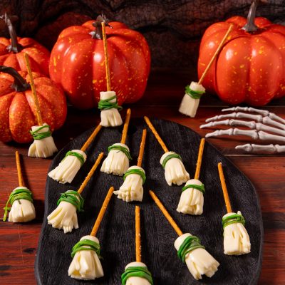 Scope delle streghe con stick di pretzel e formaggio sfilacciato legato da erba cipollina, disposte su un tagliere scuro