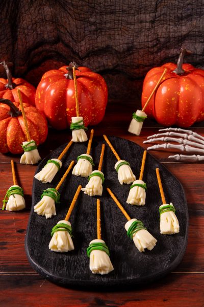 Scope delle streghe con stick di pretzel e formaggio sfilacciato legato da erba cipollina, disposte su un tagliere scuro