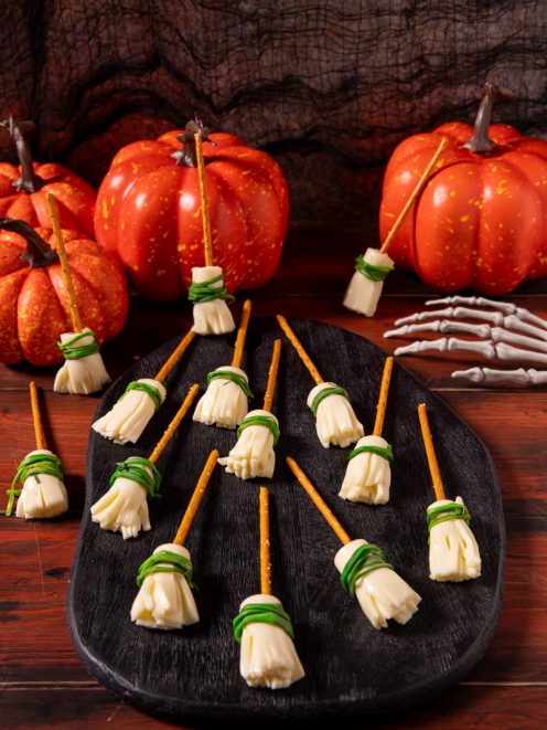 Scope delle streghe con stick di pretzel e formaggio sfilacciato legato da erba cipollina, disposte su un tagliere scuro
