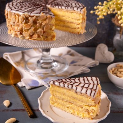 Torta Esterhazy a strati con crema gialla, glassa bianca decorata a ragnatela di cioccolato e mandorle a lamelle, con una fetta servita