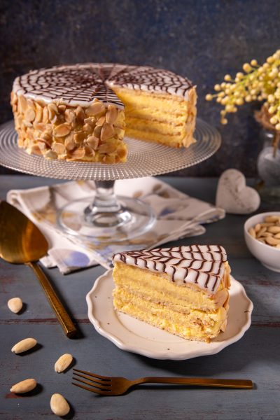 Torta Esterhazy a strati con crema gialla, glassa bianca decorata a ragnatela di cioccolato e mandorle a lamelle, con una fetta servita