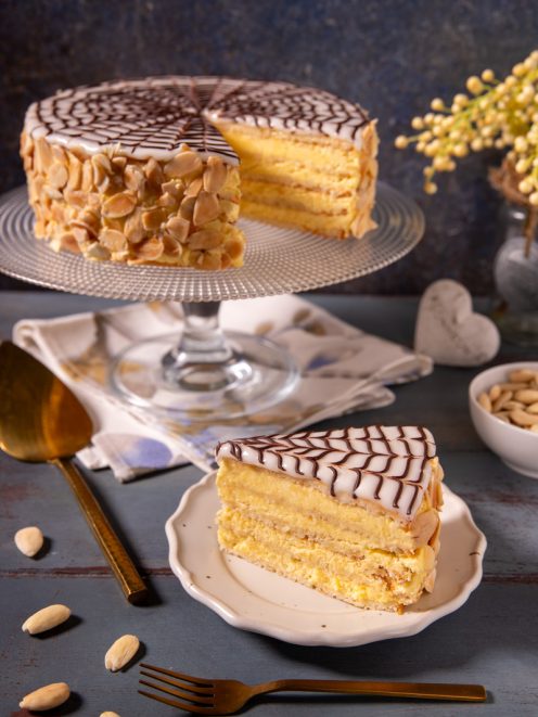 Torta Esterhazy a strati con crema gialla, glassa bianca decorata a ragnatela di cioccolato e mandorle a lamelle, con una fetta servita